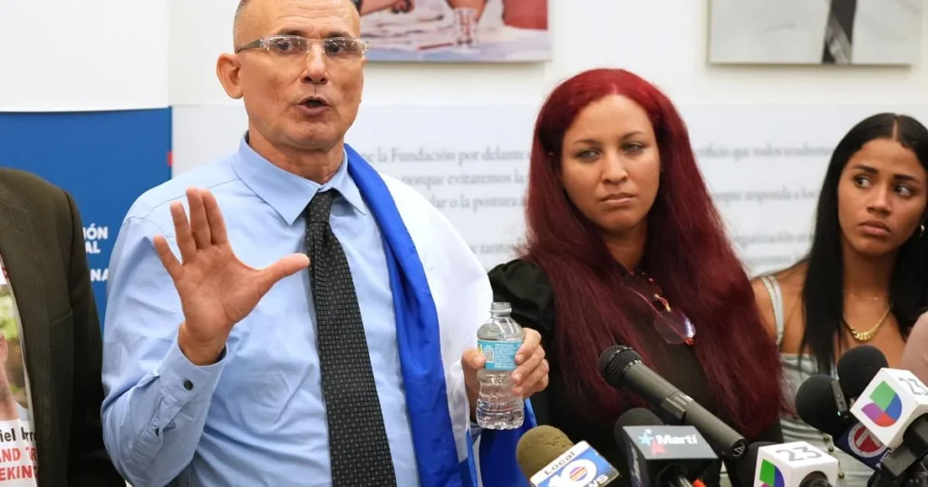 José Daniel Ferrer se exilia en Estados Unidos tras años de persecución en Cuba A man in a blue shirt and tie holds a water bottle while gesturing, surrounded by women at a press conference with microphones.