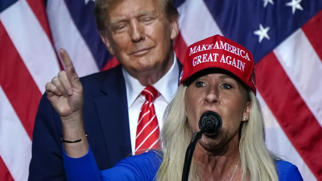 A woman wearing a red "Make America Great Again" hat raises her hand at a political event, with American flags in the background.