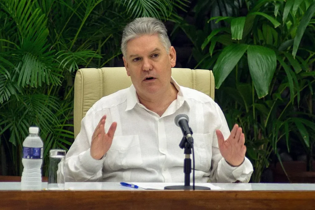 A person in a white shirt speaking at a microphone sits at a table with a water bottle and glass. Lush green plants are in the background.