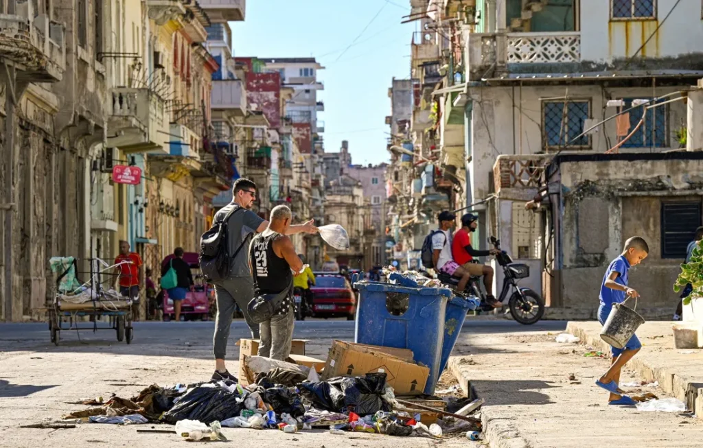 Street scene with people near overflowing blue trash bin and scattered garbage in an urban area with old buildings and motorcycles.
