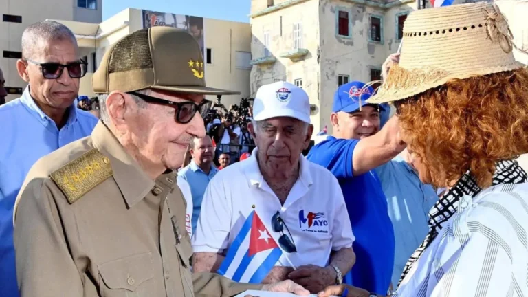 Group of people, one in military uniform, others in casual wear, interact outdoors near aged buildings under clear sky.